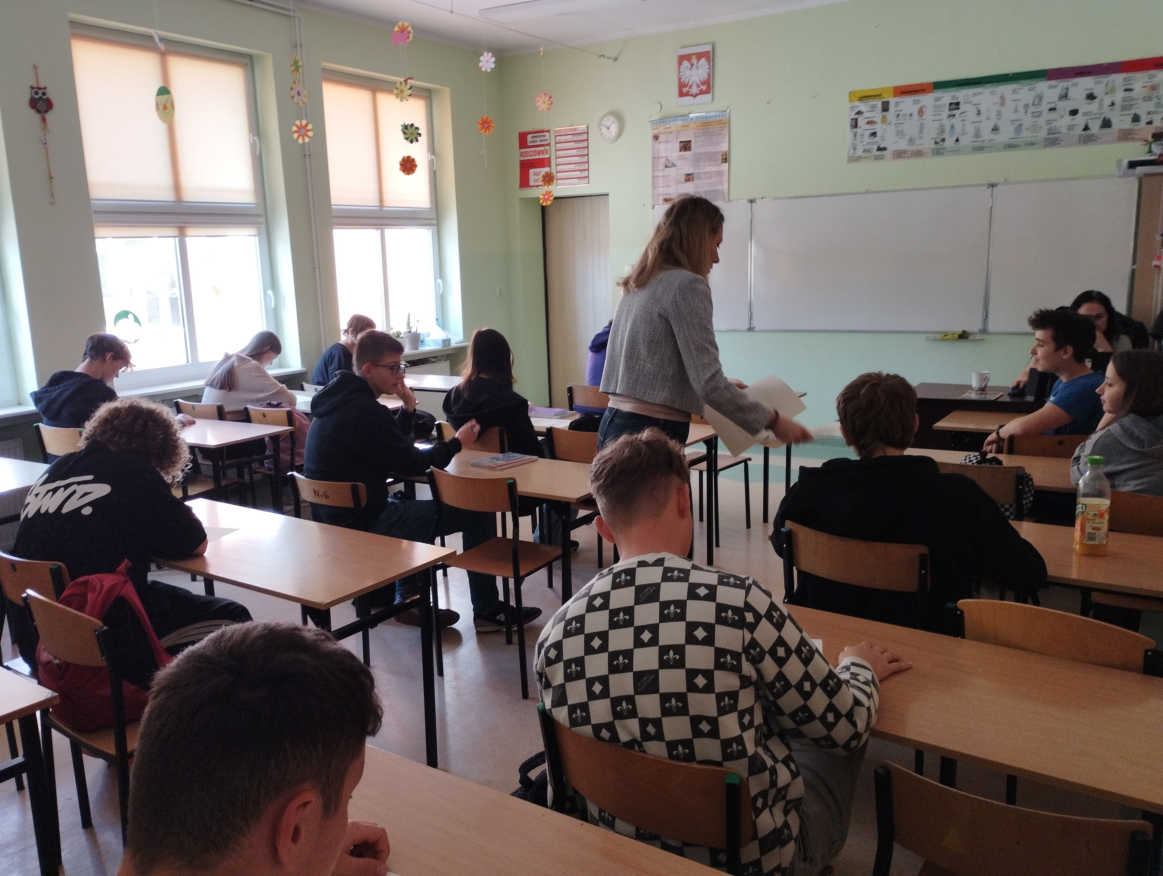 Zdjęcie przedstawia uczniów pracujących nad zadaniem. Koordynatorka rozdaje młodzieży pomocne arkusze ćwiczeniowe 