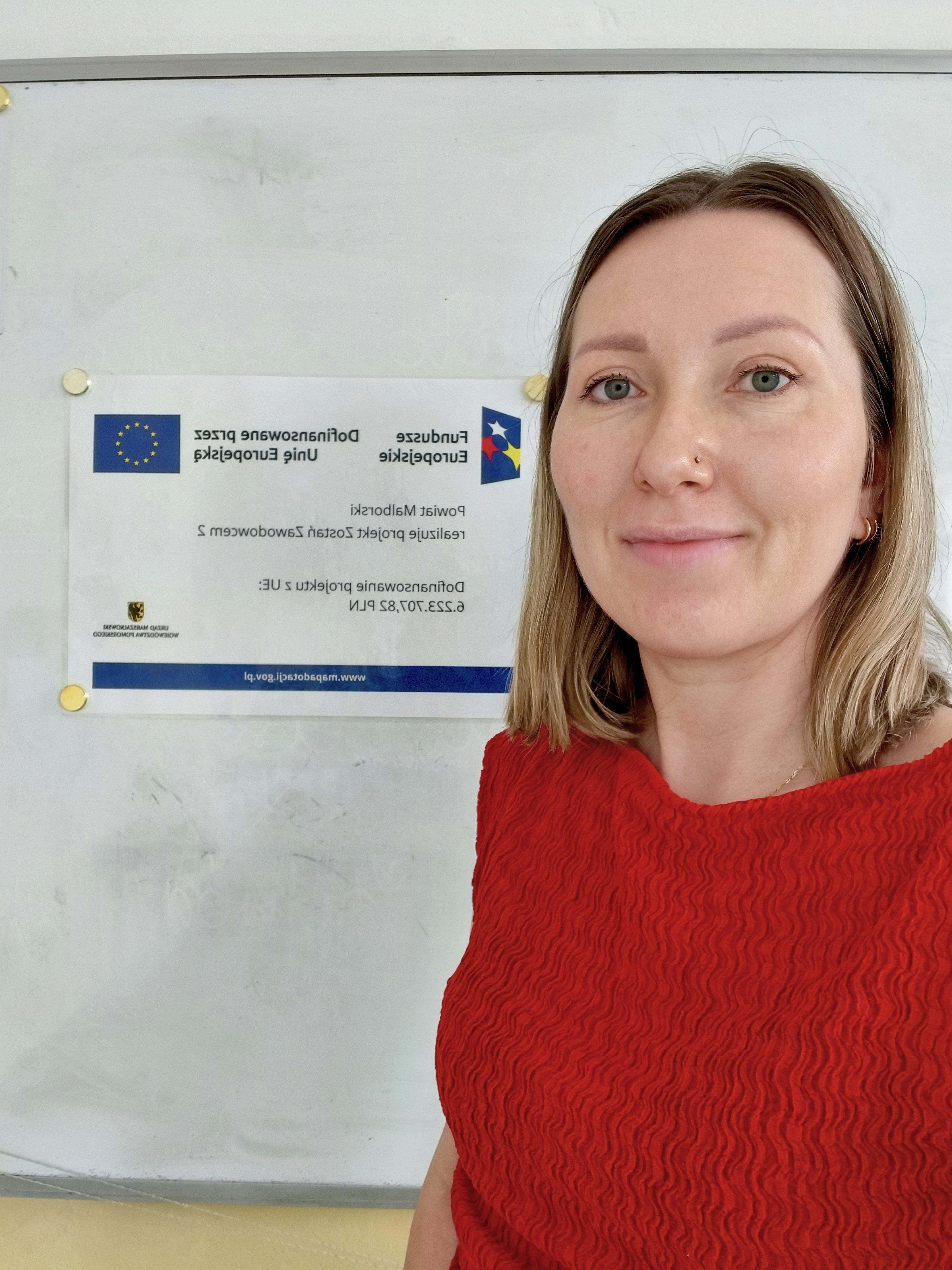 Zdjęcie przedstawia Koordynatorkę Powiatową stojącą przy tablicy, na której wisi plakat informujący o realizacji projektu "Zostań Zawodowcem 2"